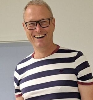 Lachender Mann mit Brille und kurz geschnittenem, blondem Haar in gestreiftem T-Shirt vor hellem Hintergrund.