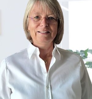 Freundlich lächelnde Frau mit kurzem, blondem Haar und Brille, trägt eine weiße Bluse und steht vor einem hellen Hintergrund mit Fensterlicht.