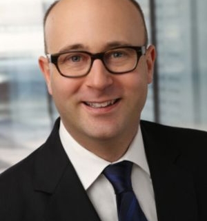 Porträt eines freundlichen Mannes mit Brille und Glatze im dunklen Anzug mit weißem Hemd und blauer Krawatte vor modernem Bürohintergrund.