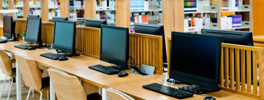 Reihe moderner Desktop-Computer mit Tastaturen und Mäusen in einem offenen Arbeitsbereich mit Holztrennwänden, umgeben von Bücherregalen.