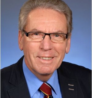 Älterer Mann mit grauen Haaren und Brille trägt Anzug, blaues Hemd und gestreifte Krawatte, vor neutralem Hintergrund.