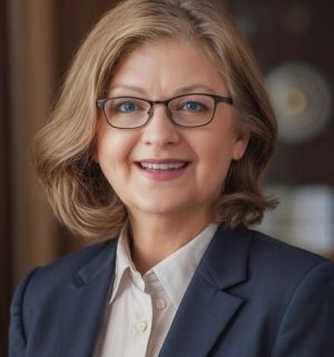 Lächelnde Frau mit schulterlangem, hellbraunem Haar, Brille und dunkelblauem Blazer vor einem warmen, unscharfen Hintergrund.