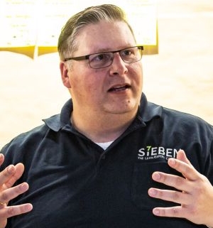 Mann mit Brille und dunkelblauem Poloshirt mit Logo „SIEBEN – The Lean Experts“ spricht gestikulierend vor einem Flipchart mit gelben Moderationskarten.