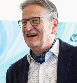 Porträt eines älteren Mannes mit Brille, grauem Haar und Halstuch, der herzlich lacht – freundliche und erfahrene Ausstrahlung vor hellem Hintergrund.