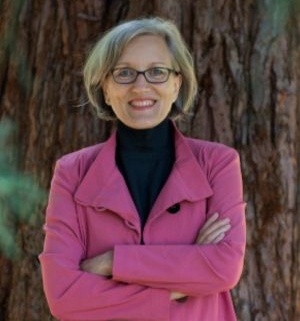 Frau mit grauem Haar und Brille steht lächelnd mit verschränkten Armen vor einem Baum. Sie trägt einen pinkfarbenen Blazer über einem schwarzen Rollkragenpullover.